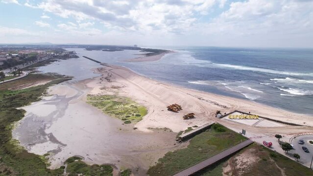 Esposende, Portugal, April 10, 2022: DRONE AERIAL FOOTAGE - The two sides of Restinga de Ofir. Esposende fishing harbor maintenance dredging. Dumper vehicles and rotating excavator parked on the sand.