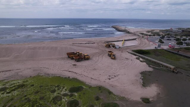 Esposende, Portugal, April 10, 2022: DRONE AERIAL FOOTAGE - The two sides of Restinga de Ofir. Esposende fishing harbor maintenance dredging. Dumper vehicles and rotating excavator parked on the sand.