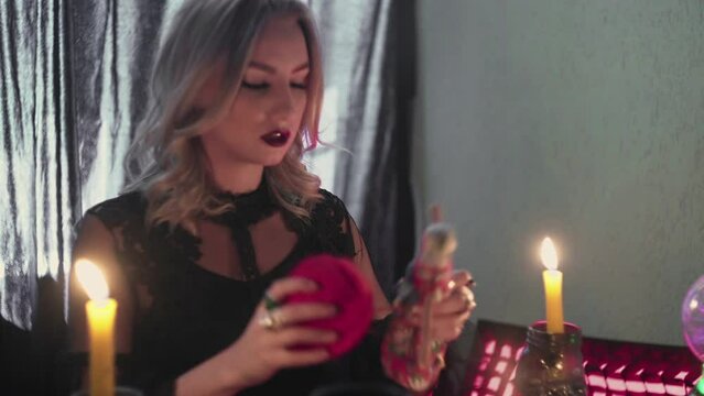 Dark magic voodoo witch performing ritual and casting spell piercing doll with needles sitting in dark room with burning candles.