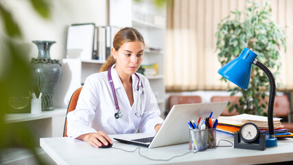 Young woman professional physician filling up medical forms on laptop while sitting at table in office.. - obrazy, fototapety, plakaty