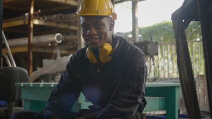 Portrait American industrial black young worker man smiling with yellow helmet in front of machine,...