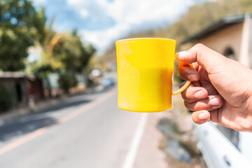 Close-up of a hand holding a plastic cup with coffee in a traditional mountainous landscape of...