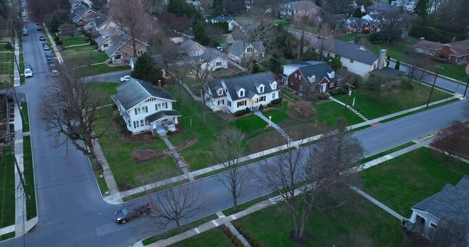 Vehicle with headlights at night drives on wide street through American residential community. Lights on at houses along road. Spring season.