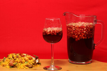 Paella rice with seafood traditional dish from Valencia Spain served on a white plate next to a pitcher of clericot with fruits and a crystal glass on a red background
