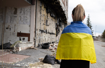 Fototapeta na wymiar Woman with Ukrainian flag looking at abandoned destroyed building. Russia invasion, war in Ukraine, crisis and destruction concept. Girl standing in front of ruined, bombed residential house.