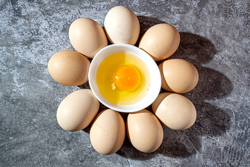 A broken raw chicken egg in a white cup surrounded by whole ones in the form of a daisy on a background of gray marble