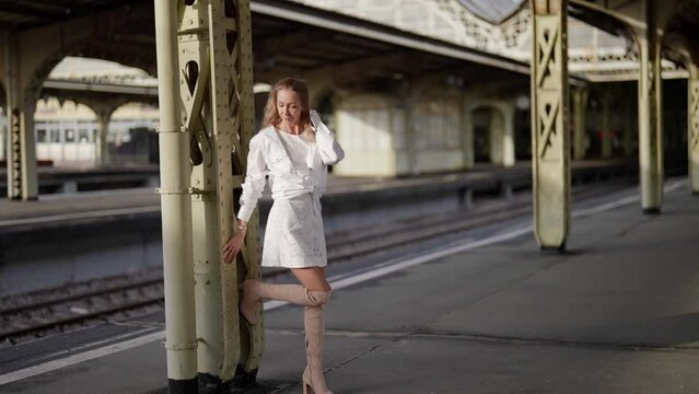 a slender pretty blonde woman in a white dress stands at an old railway station