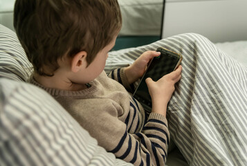Adorable boy playing online game with a smart phone. Boy playing on a smart phone. Casual child touching a mobile phone on bed. Little child with smartphone in room. Danger of internet.