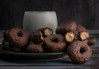 A dark plate of chocolate donuts with two biten donuts on top, and a glas of ice milk over a wooden table with a dark blue background, dark food photography