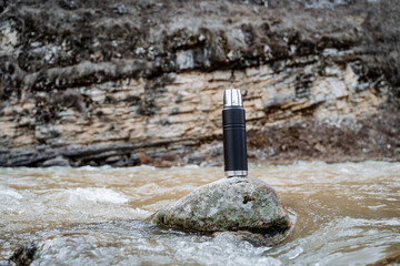 The concept of a metal vacuum thermos against the background of nature, a thermo bottle stands on a stone in the river, Hiking spring rocks. Tourist utensils.