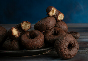 A dark plate of chocolate donuts with two biten donuts on top, over a wooden table with a dark blue background dark food photography