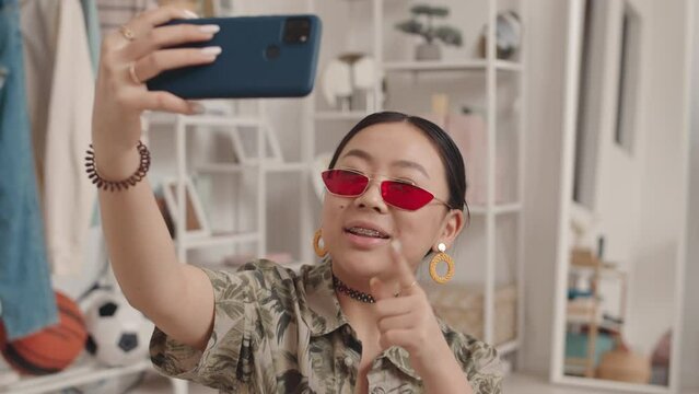 Medium closeup with slowmo of cheerful Asian gen z girl with braces in trendy clothes and accessories making live stream on smartphone from home