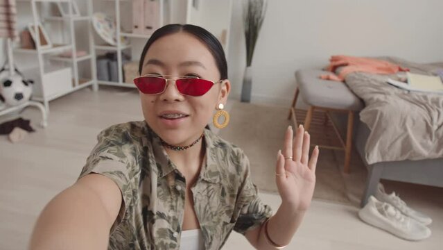 Handheld POV slowmo of Asian gen z girl in trendy shades recording video blog or making live stream from home