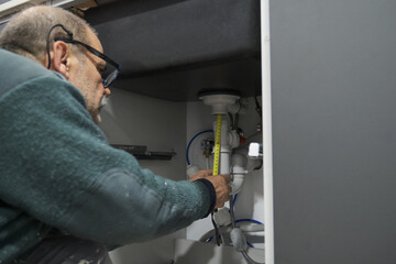 Mature plumber measuring the pipe length to install sink siphon in domestic kitchen. Repairman working.
