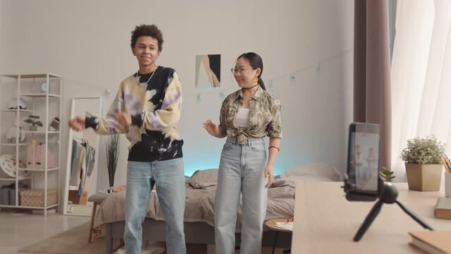 Slowmo of two cheerful diverse zoomers in blue jeans having fun while recording dance video tutorials on smartphone standing on tripod on desk, spending leisure time together at home
