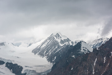Atmospheric landscape with sharp rocks and high snowy mountain top in rainy low clouds at overcast. Dramatic gloomy scenery with large snow mountains and glacier in gray cloudy sky at rainy weather.