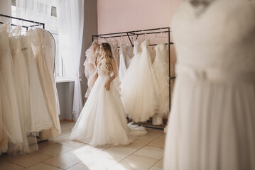 Young blonde woman trying on a dress in wedding salon. A bride with wedding dresses and mannequins in boutique. Full length portrait - Powered by Adobe