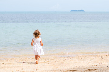 Fototapeta na wymiar A little girl with curly hair in a white dress walks along a sandy beach on the shore of the sea, ocean and smiles. Sea holidays, travel and beach holidays with children