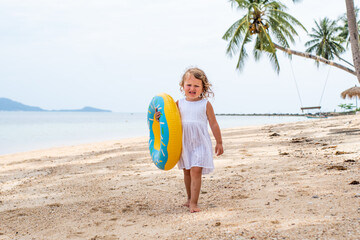 Naklejka na ściany i meble A little girl with curly hair in a white dress walks along a sandy beach on the shore of the sea, ocean and smiles. cute girl with an inflatable swimming circle