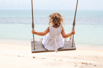 A little girl with curly hair in a white dress is swinging on a swing on a sandy beach by the sea, ocean. Sea holidays, travel and beach holidays with children