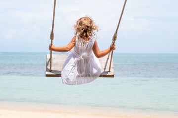 A little girl with curly hair in a white dress is swinging on a swing on a sandy beach by the sea, ocean. Sea holidays, travel and beach holidays with children