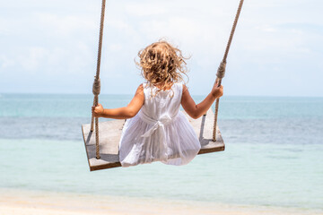 A little girl with curly hair in a white dress is swinging on a swing on a sandy beach by the sea, ocean. Sea holidays, travel and beach holidays with children