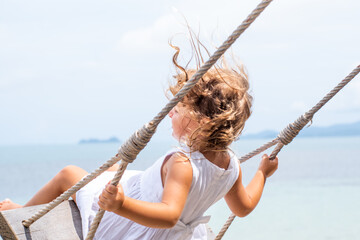 A little girl with curly hair in a white dress is swinging on a swing on a sandy beach by the sea, ocean. Sea holidays, travel and beach holidays with children