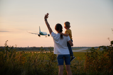 Back view of mother with toddler waving hands to landing commercial airplane at sunset. Lifestyle...