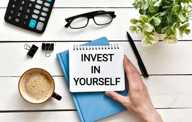 Female hand holding a notebook with the text Invest in yourself on a diary on white wooden table with eyeglasses, green plant, calculator and pen