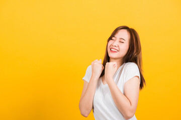 Asian happy portrait beautiful cute young woman teen standing wear t-shirt makes raised fists up celebrating her success looking to camera isolated, studio shot on yellow background with copy space