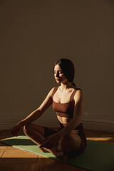 Peaceful young caucasian woman sitting on green yoga mat practicing meditation in lotus position. Brunette with collected hair wears top and leggings. Concept of relaxation and rest