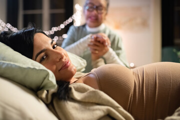 Smiling pregnant woman and midwife at home. Woman in casual clothes lying on bed, Asian doula holding hand. Pregnancy, medicine, home birth concept - obrazy, fototapety, plakaty