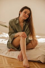 Attractive young caucasian woman sitting barefoot on mattress looking at camera with carefree smile. Brunette woman with long hair wears casual clothes. Leisure, comfort and people