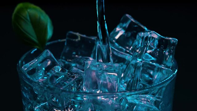 Static shot of artistic cocktail with blue light and coloring being poured over ice with a black background and a green leaf garnish