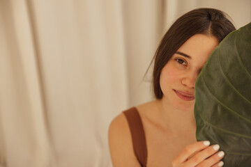 Obraz na płótnie Canvas Tender young caucasian woman with brown eyes covered her face with green tropical leaf. Brunette with loose hair wears open top. Good health, healthy lifestyle concept