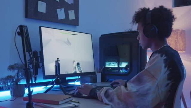 Waist up slowmo on 14 year old Biracial boy in headphones making live stream on smartphone while playing fps game on pc at home