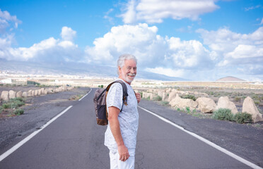 Smiling alone traveler senior man walking outdoors enjoying freedom looking at camera. Elderly mature man in empty road, mountain and blue sky on background