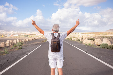Fototapeta na wymiar Back view of senior caucasian man with backpack on shoulders walking in empty street outdoors gesturing peace or victory with open arms. White-haired old man enjoying freedom and nature. Copy space