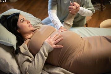 Cropped image of pregnant woman and caring midwife at home. Woman in casual clothes lying on bed,...