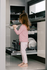 Cute toddler girl wearing casual clothes, holding clean plates. Kid helping in the kitchen.