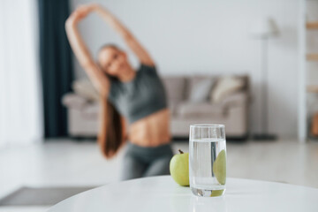 With glass of water. Young woman with slim body type and in yoga clothes is at home