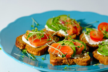toasts with salmon, cottage cheese, avocado, microgreens, cherry tomatoes in blue plate on white background, open fish sandwiches