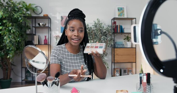 Smiling brunette female beauty blogger recommends products showing eye shadow palette. She records video using smartphone and ring light. Female makeup artist influencer creates tutorial.