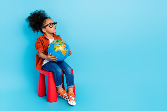 Full size photo of interested little girl casual style look empty space thinking hold globe isolated on blue color background
