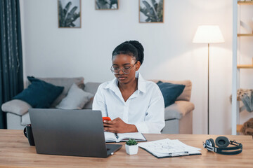 Fototapeta premium Working by using laptop. Young african american woman in white shirt is at home