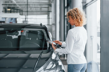 Holding tablet in hands. Woman with curly blonde hair is in autosalon