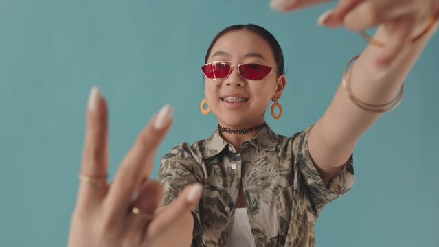 Waist up slowmo portrait of happy Asian teenage girl in trendy eyeglasses showing k-pop inspired hand signs at camera and smiling with her braces standing on turquoise background