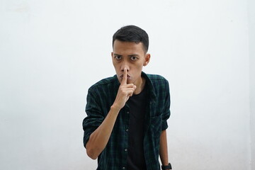 Fototapeta na wymiar Serious asian man wearing dark green casual wear holding finger near lips, showing shh gesture asking to keep quiet and still, privacy. Indoor studio shot isolated on a white background