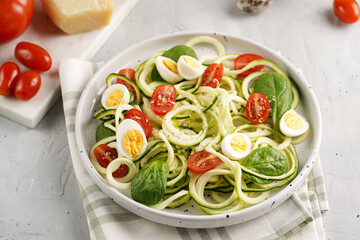 Keto paleo low-carb zoodles: zucchini noodles with parmesan cheese, cherry tomatoes, quail eggs on a checkered kitchen towel light grey surface