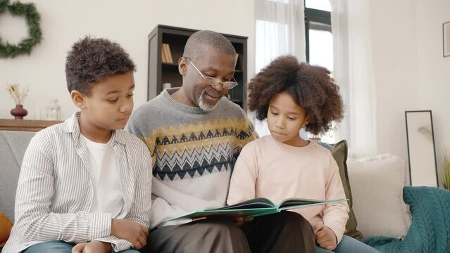 Happy family life. Caring african american grandfather in eyeglasses reading book to cute boy and girl, free space